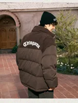 Campera puffer marrón con paneles horizontales acolchados. La parte superior de la espalda es de pana marrón con el texto 'Chingona' bordado en blanco. Posee cordón ajustable en el ruedo y se lleva superpuesta a una sudadera con capucha gris.