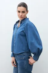 Camisa de jean de color azul medio, con cuello camisero, botones dorados y mangas largas abullonadas con puños ajustados.