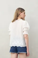 Blusa azul grisáceo de algodón con bordados frontales y terminaciones festoneadas. Tiene mangas cortas con frunces y cuello redondo con detalle de puntilla.