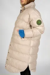Campera puffer larga color beige, con cuello alto y cierre frontal con botones a presión. Tiene bolsillos laterales y un parche con el logo de la marca en la manga izquierda.