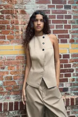 Top chaleco color beige de cuello halter con espalda descubierta y trabilla con hebilla metálica. Cierre frontal con botones y ganchos metálicos.