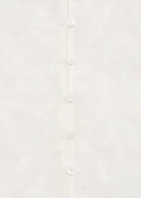 Blusa blanca de encaje floral semitransparente, con cuello redondo, mangas largas y cierre de botones en la nuca.