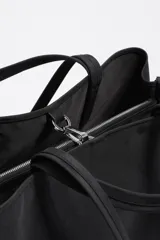 Bolso shopper grande de nylon negro, con estructura blanda, doble asa de mano y correa larga ajustable para llevar al hombro. Presenta un logo metálico con el texto "BIMBA Y LOLA" en la parte frontal inferior.