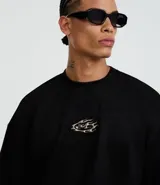 Remera de manga corta, cuello redondo y corte boxy, confeccionada en gamuza sintética (suede) de color negro. Presenta una pequeña estampa localizada en el pecho con un diseño abstracto en tonos dorados/bronce.