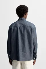Camisa azul de algodón con textura chambray, corte regular fit, cuello abotonado, manga larga con puños abotonados, bolsillo de parche en el pecho y cierre frontal con botones.
