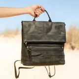 Mochila verde de símil cuero con tapa y broches magnéticos, bolsillo frontal, interior con división y bolsillos internos. Correas desmontables que permiten transformarla en cartera.