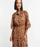 Vestido largo de chiffon con estampa de corazones con diseño de leopardo. Tiene cuello alto con lazo, mangas largas ligeramente transparentes y falda con volado. El tejido contiene hilo metalizado.