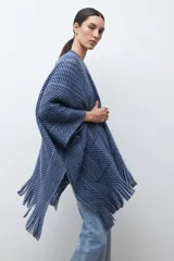 Ruana de tejido de punto en tonos beige y blanco, con patrón de espiga. Presenta cuello en V, mangas amplias, dos bolsillos frontales y flecos en el dobladillo.