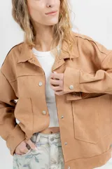 Chaqueta color beige de corte recto, estilo camisa, confeccionada en gamuza con elastano. Presenta cuello camisero, cierre frontal con botones metálicos, bolsillos grandes en el frente y charreteras en los hombros.