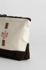 Neceser de lona color crudo con base y cierre superior en marrón oscuro. Estampado frontal con el texto "The Beach Club" en rojo y dos líneas verticales con franjas rojas y blancas.