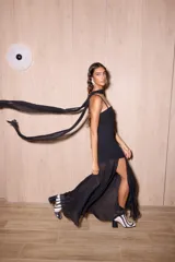 Vestido negro de largo midi, con tirantes finos y escote recto. La falda es asimétrica, con una capa superior de gasa transparente que cae hasta el tobillo y una base más corta de crepé elastizado. Incluye una bufanda larga de gasa a juego que se ata al cuello.