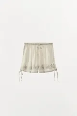 Shorts de tiro alto color crudo, con cintura elástica ajustable con lazada. Detalle de bordados florales combinados a tono, aplicación de puntilla y abalorios. Bajo acabado con tira ajustable.