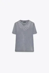 Camiseta gris de algodón con cuello redondo y mangas cortas. Presenta apliques de strass plateados alrededor del cuello en forma de V.