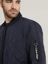 Campera bomber azul marino con diseño acolchado en forma de rombos, cierre frontal con cremallera, cuello, puños y dobladillo acanalados, bolsillos laterales y bolsillo en la manga con cremallera. Logotipo de Calvin Klein en la manga.