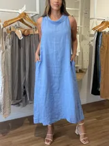 Vestido largo sin mangas color azul claro, confeccionado en lino, con cuello tipo buche o drapeado. Presenta un calce holgado y bolsillos laterales.