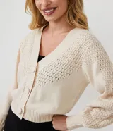 Cardigan corto de punto color crema, con escote en V, abotonadura frontal y mangas largas abullonadas con puños acanalados. Presenta un punto calado en forma de rombos en la parte superior del busto y en las mangas.