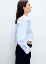 Camisa corta de popelina de algodón a rayas verticales celestes y blancas, con cuello camisero, mangas largas con puños abotonados, bolsillo de parche en el pecho y cierre frontal con botones.