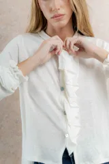 Blusa color crudo confeccionada en lino, con volados y botones en la delantera, cuello a la base y manga larga con elástico en los puños.