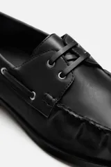 Zapato náutico de vestir color negro, con parte superior lisa, bordón, detalle de ojales metálicos, horma redonda y suela a tono con ligero tacón.