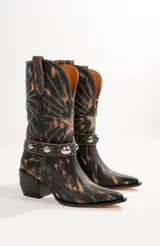 Botas texanas de cuero negro con detalles desgastados en color ocre y bordados en hilo blanco. Presentan una correa de cuero con tachas plateadas alrededor del tobillo y un aplique metálico en forma de estrella.
