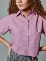 Camisa corta de lino color lila con cuello clásico, cierre frontal con botones, mangas cortas y dos bolsillos de parche en el pecho.
