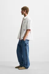 Camisa blanca de algodón con estructura canvas, corte boxy fit, cuello bowling y manga corta. Presenta bordado en azul claro con texto cursivo en el pecho y botones frontales.