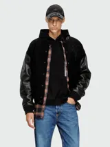 Campera estilo universitaria (varsity jacket) negra con mangas de cuero sintético negro y cuerpo de lana negra. Presenta un parche bordado grande en el pecho y cierre frontal de botones a presión. Tiene cuello, puños y cintura acanalados.