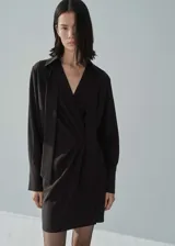 Vestido corto negro con cuello camisero, escote en V cruzado, mangas largas con puños abotonados, lazo desmontable en el cuello y detalle de pliegues en la cintura.