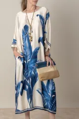 Vestido largo color natural con estampado de hojas de palmera en tonos verdes. Tiene corte amplio tipo túnica, cuello redondo, mangas tres cuartos, bolsillos laterales y ruedo con ribete.