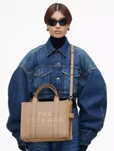 Bolso tote mediano de cuero color beige, con asas superiores y correa ajustable y desmontable para llevar al hombro o cruzado. Presenta el logo en relieve en el frente: "THE TOTE BAG MARC JACOBS".