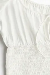 Blusa blanca de viscosa con escote cuadrado, mangas cortas abullonadas con elástico en hombros y puños, cintura fruncida y abertura con cordón en el delantero.