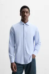 Camisa celeste a rayas verticales blancas, de corte recto, con cuello italiano abierto, manga larga con puños abotonados y cierre frontal con botones.