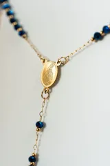 Collar estilo rosario con cadena dorada y cuentas pequeñas de color azul oscuro. Incluye un dije ovalado (medalla) y una cruz colgante en el extremo inferior.