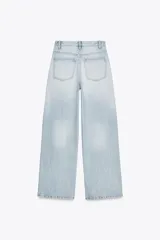 Pantalón de jean celeste de tiro alto, corte wide leg y largo hasta el tobillo. Presenta un diseño asimétrico en la cintura con cierre frontal cruzado con cremallera y botones metálicos.