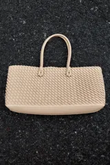 Bolso shopper alargado color beige con diseño trenzado, doble asa de hombro y cierre con botón imantado. Incluye bolsa interior con cierre fruncido.
