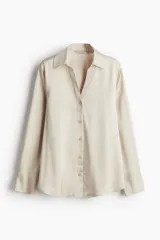 Camisa color crema de tela satinada, con escote en V, cuello clásico, cierre frontal con botones, mangas largas con puños abotonados y ruedo redondeado.
