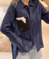 Camisa color naranja de lino o algodón con cuello clásico, cierre frontal con botones y mangas largas con puños abotonados. Presenta un bolsillo de cuero marrón en el pecho.