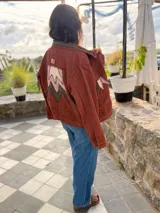 Campera bomber de jean color terracota, con corte holgado. Presenta apliques de cuero cortados y bordados a mano en la espalda, delantera y cuello, formando un diseño de montañas en tonos beige, rosa y verde militar.