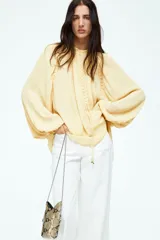 Blusa color crema de gasa transparente con cuello elevado y mangas globo largas con puños elásticos. Presenta pespuntes sobredimensionados decorativos en las costuras raglán y tira diagonal adelante con colgantes de cuentas brillantes.