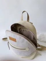 Mochila pequeña de cuero color gris claro y beige, con cierre superior y bolsillo frontal con cierre. Tiene asa de mano y correas ajustables para los hombros.
