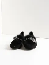 Zapato plano negro estilo mocasín con detalle de pelo sintético negro en la parte delantera y una tira de cuero sintético con hebilla metálica plateada y ojales. El talón es bajo y de material liso.