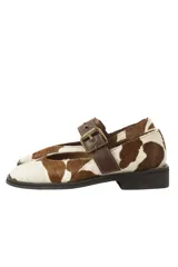 Zapatos Mary Jane con estampado de vaca en blanco y marrón, correa de cuero marrón con hebilla dorada y suela plana de goma negra.