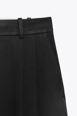 Pantalón negro de tiro alto con trabillas, bolsillos laterales y pinzas. Cierre frontal con cremallera, botón interior y gancho metálico.