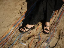 Sandalias negras de plataforma baja con dos tiras finas adornadas con rosetas negras. Una tira cruza sobre los dedos y la otra funciona como pulsera tobillera.