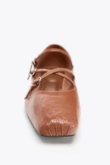 Zapato tipo bailarina color marrón de piel con acabado charol. Tiene punta cuadrada, detalle de tiras con hebillas metálicas y suela plana.