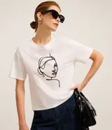 Remera blanca de algodón con cuello redondo y bordado de una cara de mujer con caravana dorada.