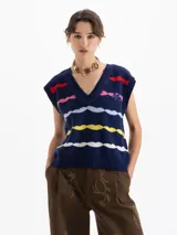 Chaleco de punto azul marino con escote en V y bordes trenzados. Presenta un patrón de ondas horizontales en colores rojo, rosa, azul claro, amarillo y blanco. Incluye un bordado tipo escudo en el frente.