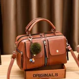 Cartera tipo baúl color marrón, con doble asa corta y correa larga desmontable. Tiene dos bolsillos frontales, uno con cierre y otro con solapa y botón. Incluye un charm colgante de pompón y detalles de costuras en contraste.