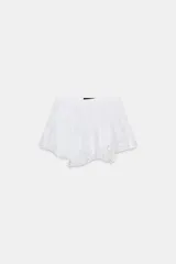 Falda corta blanca de tiro medio con bordados perforados (ojales) y bajo con volantes. Presenta un detalle de lazo ajustable en un lateral de la cintura.