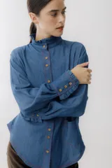 Camisa celeste de jean con cuello alto con volados y mangas largas abullonadas con puños con volados. Presenta botones de madera y costuras rojas a contraste.